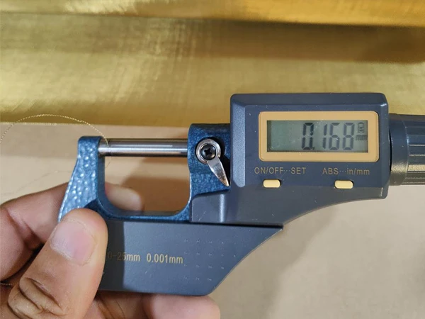 A person holding a micrometer measuring the wire diameter of the woven brass wire mesh screen as 0.168 mm